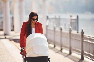 pratend door de telefoon. moeder in rood jas hebben een wandelen met haar kind in de kinderwagen in de park Bij herfst tijd foto