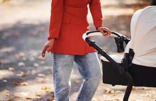 houdt sigaret in hand. moeder in rood jas en met sigaret hebben een wandelen met haar kind in de kinderwagen in de park Bij herfst tijd foto