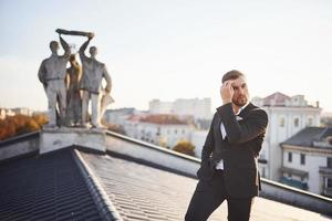 Mens in zwart luxe pak staat Aan de op het dak tegen standbeeld foto
