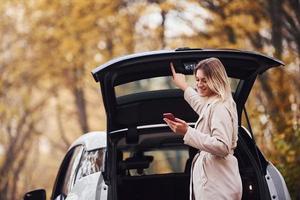 gebruik makend van smartphone. meisje hebben herfst reis door auto. modern merk nieuw auto- in de Woud foto