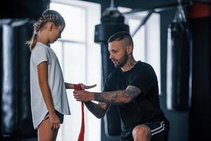 helpen naar slijtage de verbanden. jong getatoeëerd boksen trainer geeft les schattig weinig meisje in de Sportschool foto