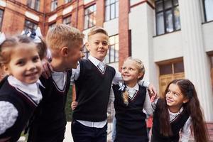 hebben pret en omarmen elk ander. groep van kinderen in school- uniform dat is buitenshuis samen in de buurt onderwijs gebouw foto