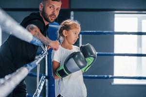 staand Aan de boksen ring. jong getatoeëerd boksen trainer geeft les schattig weinig meisje in de Sportschool foto
