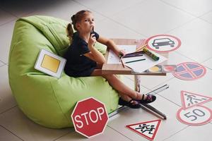 schattig weinig meisje zit Aan de zacht groen stoel door de tafel met potlood en papier bladen. in de buurt modern auto- en weg tekens Aan de verdieping foto