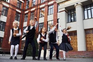 groep van kinderen in school- uniform jumping en hebben pret buitenshuis samen in de buurt onderwijs gebouw foto