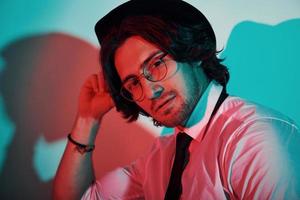 portret van jong elegant Mens in hoed en met bril dat staat in neon lichten in de studio foto