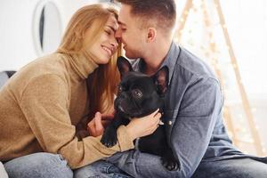 gelukkig jong modern paar zittend Aan de sofa Bij huis met Kerstmis boom met hun schattig hond foto