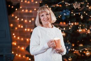 senior vrolijk vrouw staand met kop van drinken in de kamer met Kerstmis decoraties foto