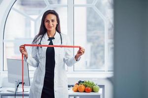 vrouw voedingsdeskundige in wit jas staand binnenshuis in de kantoor Bij werkplaats met meten plakband in handen foto