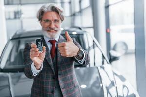 senior zakenman in pak en stropdas met grijs haar- en baard staand binnenshuis in de buurt auto met sleutels in hand- foto