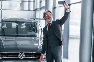 positief senior Mens met grijs haar- nemen selfie tegen modern auto binnenshuis foto