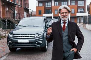 modieus senior Mens met grijs haar- en baard staand buitenshuis Aan de straat in de buurt zijn auto met sleutels in hand- foto