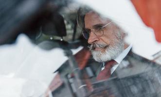 visie door de glas van elegant senior Mens met grijs haar- en snor dat is in de modern auto foto
