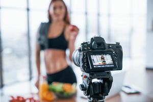 vrouw vlogger met sportief lichaam staand binnenshuis in de buurt tafel met gezond voedsel foto