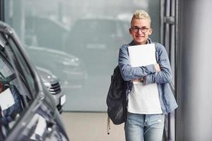 jong hipster vent in mooi hoor kleren staat binnenshuis tegen grijs achtergrond en in de buurt auto foto