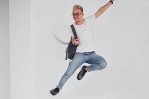 gelukkig hipster vent in gewoontjes kleren en met rugzak jumping binnenshuis tegen wit muur foto