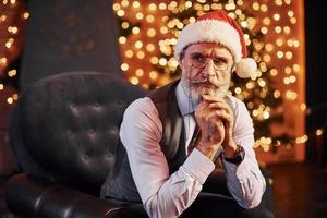 portret van elegant senior met grijs haar- en baard zittend in versierd kamer en in Kerstmis hoed foto