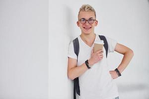 leerling in gewoontjes kleren en met rugzak staat binnenshuis tegen wit muur met kop van drinken foto