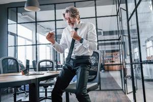 gelukkig dansen volwassen zakenman met grijs haar- en baard in formeel kleren vieren succes in de kantoor foto