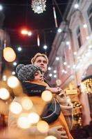 positief jong paar in warm kleren omarmen elk andere Aan Kerstmis versierd straat foto