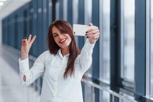 brunette in wit overhemd maken selfie binnenshuis in modern luchthaven of gang Bij dag foto