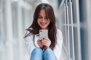 brunette in wit overhemd met telefoon in handen zit binnenshuis in modern luchthaven of gang Bij dag foto