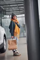 mooi vrolijk meisje in geel sjaal en in warm kleren staand binnenshuis met boodschappen doen Tassen in handen in de buurt auto foto