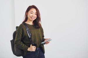 portret van gelukkig Aziatisch jong meisje met rugzak en telefoon dat staand binnenshuis in de studio tegen wit achtergrond foto