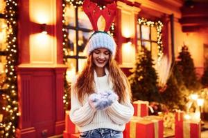 jong meisje in feestelijk hert oren Aan hoofd houdt weinig geschenk doos buitenshuis in de stad Bij avond tijd in de buurt gebouw met kunstmatig feestelijk lichten foto