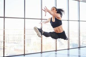 jong sportief vrouw in sportkleding jumping en aan het doen atletisch trucs tegen venster in de lucht foto