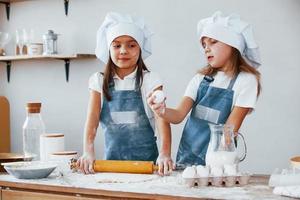 twee weinig meisjes in blauw chef uniform kneden deeg Aan de keuken foto