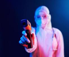 verpleegster in masker en wit uniform staand in neon verlicht kamer en Holding infrarood thermometer. hou op coronavirus verspreiden foto