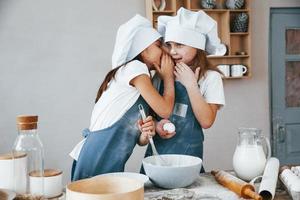 twee weinig meisjes in blauw chef uniform pratend geheimen naar elk andere wanneer voorbereidingen treffen voedsel Aan de keuken foto