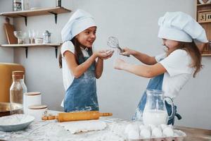 twee weinig meisjes in blauw chef uniform hebben pret en voorbereidingen treffen voedsel Aan de keuken foto