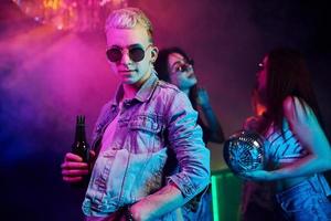 hipster vent in zonnebril en met fles van alcohol poseren voor camera in voorkant van jong mensen dat hebben pret in nacht club met kleurrijk laser lichten foto