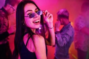 jong meisje Holding drug pillen Aan togue in voorkant van jong mensen dat hebben pret in nacht club met kleurrijk laser lichten foto