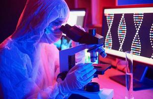 verpleegster in masker en wit uniform zittend in neon verlicht laboratorium met computer, microscoop en medisch uitrusting zoeken voor coronavirus vaccin foto