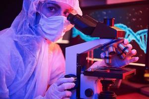 verpleegster in masker en wit uniform zittend in neon verlicht laboratorium met computer, microscoop en medisch uitrusting zoeken voor coronavirus vaccin foto