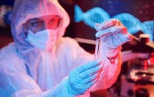 verpleegster in masker en wit uniform, Holding buis met vloeistof en zittend in neon verlicht laboratorium met computer en medisch uitrusting zoeken voor coronavirus vaccin foto