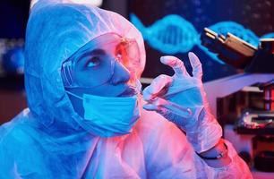 verpleegster in masker en wit uniform, Holding buis met vloeistof en zittend in neon verlicht laboratorium met computer en medisch uitrusting zoeken voor coronavirus vaccin foto