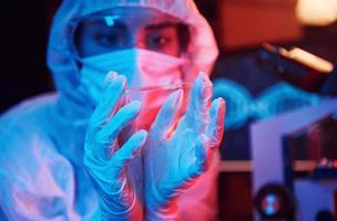 verpleegster in masker en wit uniform, Holding buis met vloeistof en zittend in neon verlicht laboratorium met computer en medisch uitrusting zoeken voor coronavirus vaccin foto