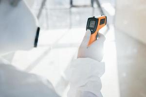 vrouw dokter wetenschapper in laboratorium jas, defensief eyewear en masker staand binnenshuis met geïnfrateerd thermometer foto