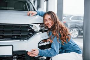 positief vrouw in blauw overhemd omarmen haar merk nieuw auto. in auto salon foto