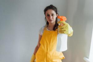 huisvrouw in geel uniform staand met schoonmaak verstuiven voor ramen binnenshuis. huis vernieuwing opvatting foto