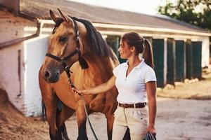 krijgen klaar voor de rijden. amazone in wit uniform met haar paard Bij boerderij. klaar voor de rijden foto