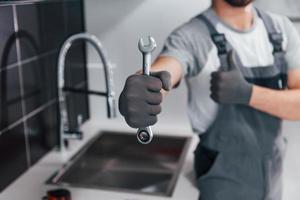 dichtbij omhoog visie van jong professioneel loodgieter in grijs uniform Holding moersleutel in hand- Aan de keuken foto