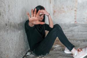 hou op de geweld. jong geslagen vrouw in zwart kleren zittend Aan de verdieping van verlaten gebouw foto