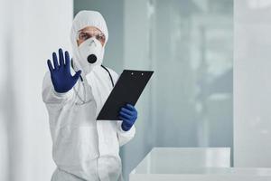 shows hou op gebaar door hand. mannetje dokter wetenschapper in laboratorium jas, defensief eyewear en masker Holding kladblok in handen foto