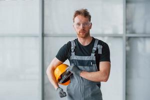 portret van Mens in grijs uniform en met oranje moeilijk hoed in handen dat staand binnenshuis in modern groot kantoor Bij dag foto