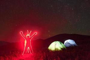 figuur van Mens door neon verlichting. twee verlicht tenten onder sterren Bij bergen Bij nacht foto